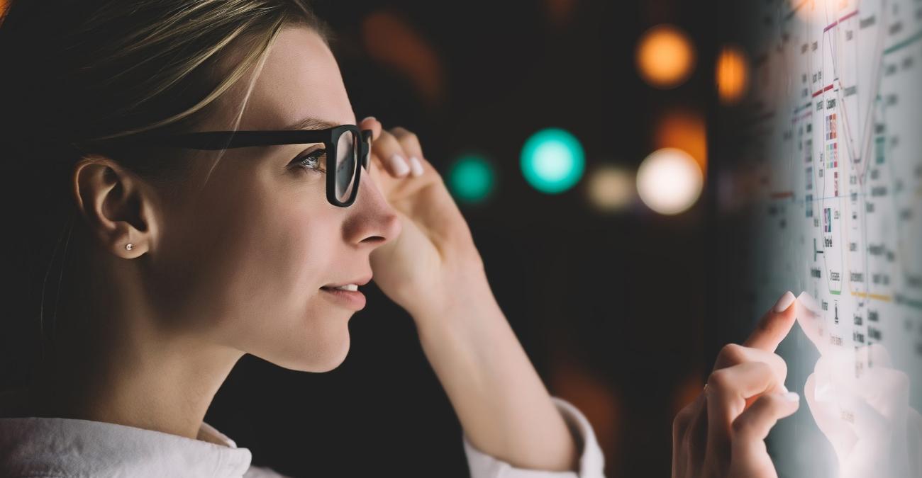 Eine Frau mit Brille betrachtet konzentriert ein hell beleuchtetes Diagramm aus zahlreichen verknüpften Punkten und Linien auf einem Touchscreen und tippt auf eine der Verbindungen, Symbolbild für API Lifecycle Management und den Überblick über vernetzte Schnittstellen.