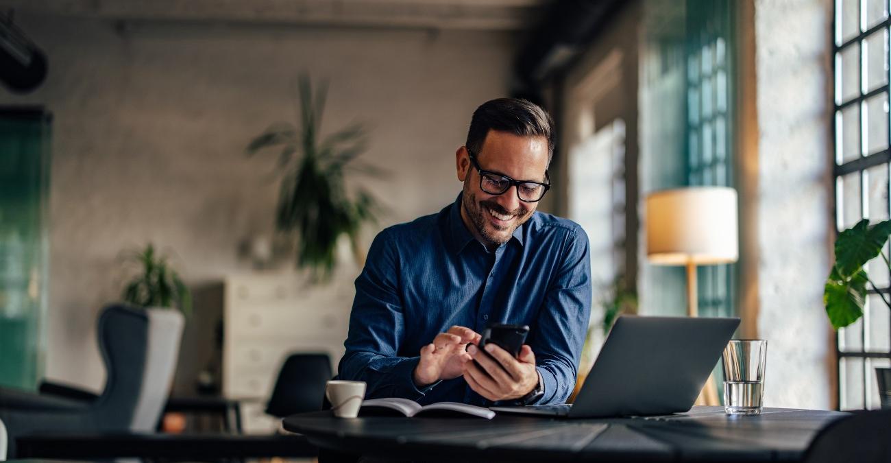 Ein helles Office mit großen Fenstern und Blumen im Hintergrund. Ein Mitarbeiter sitzt gut gelaunt am Schreibtisch vor seinem Notebook. Er nutzt seine Business-App mit ERP-Anbindung.