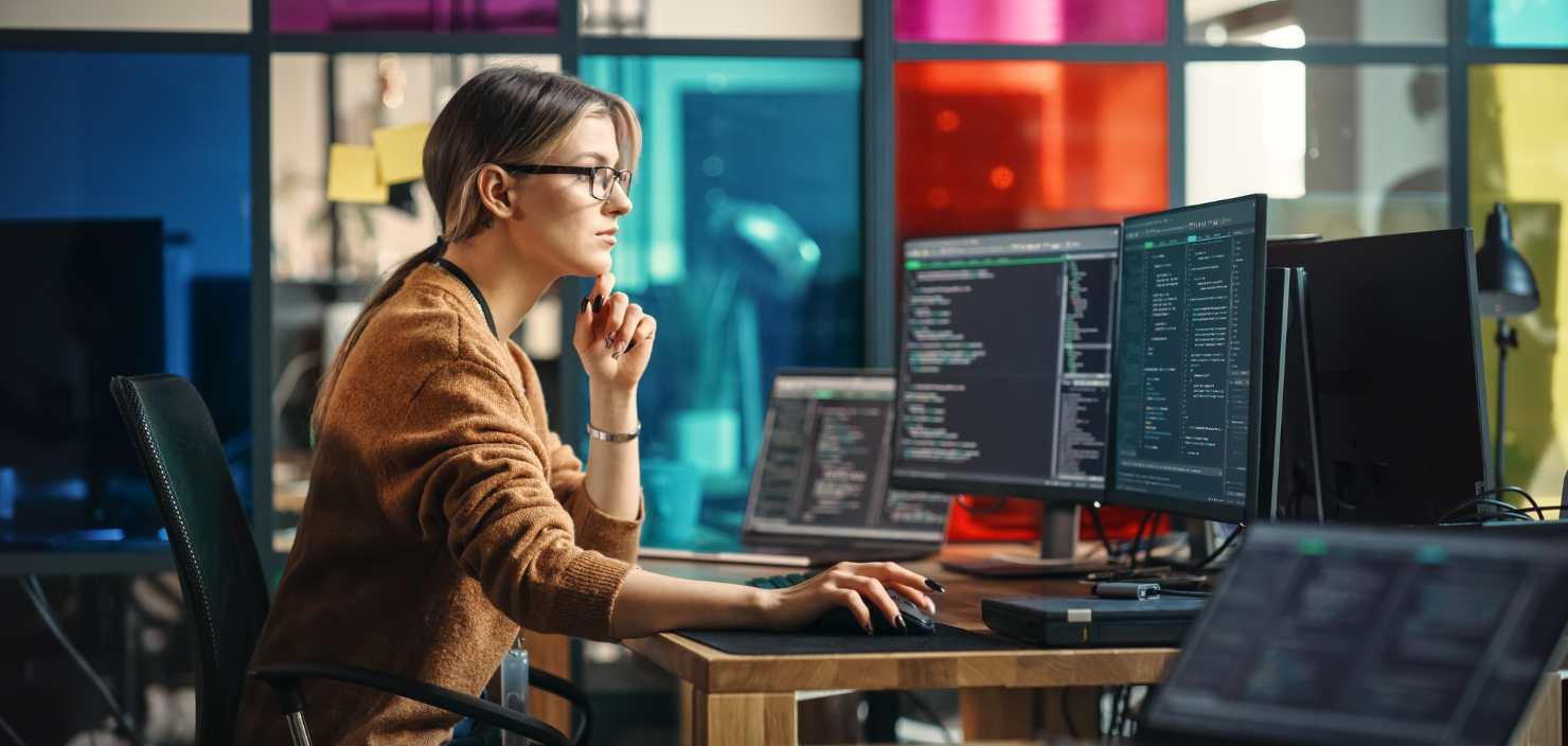 Eine junge Informatikerin sitzt an mehreren großen Bildschirmen. Sie blickt konzentriert auf die Bildschirme. Sie erfasst Prozessdaten für das Application Performance Management.