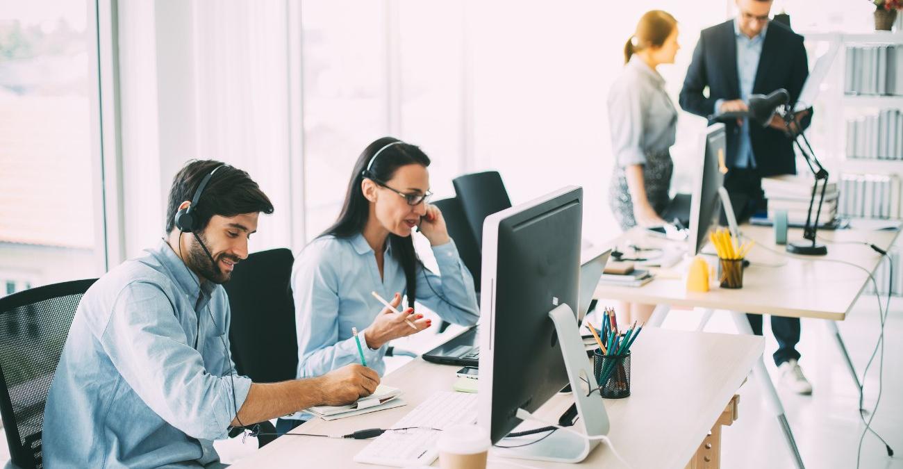 Ein freundliches Team mit Headsets arbeitet gemeinsam am Computer in einem modernen Büro – Sinnbild für den betreuten Service-Betrieb hinter Backup as a Service, bei dem ein Dienstleister die Datensicherung übernimmt.