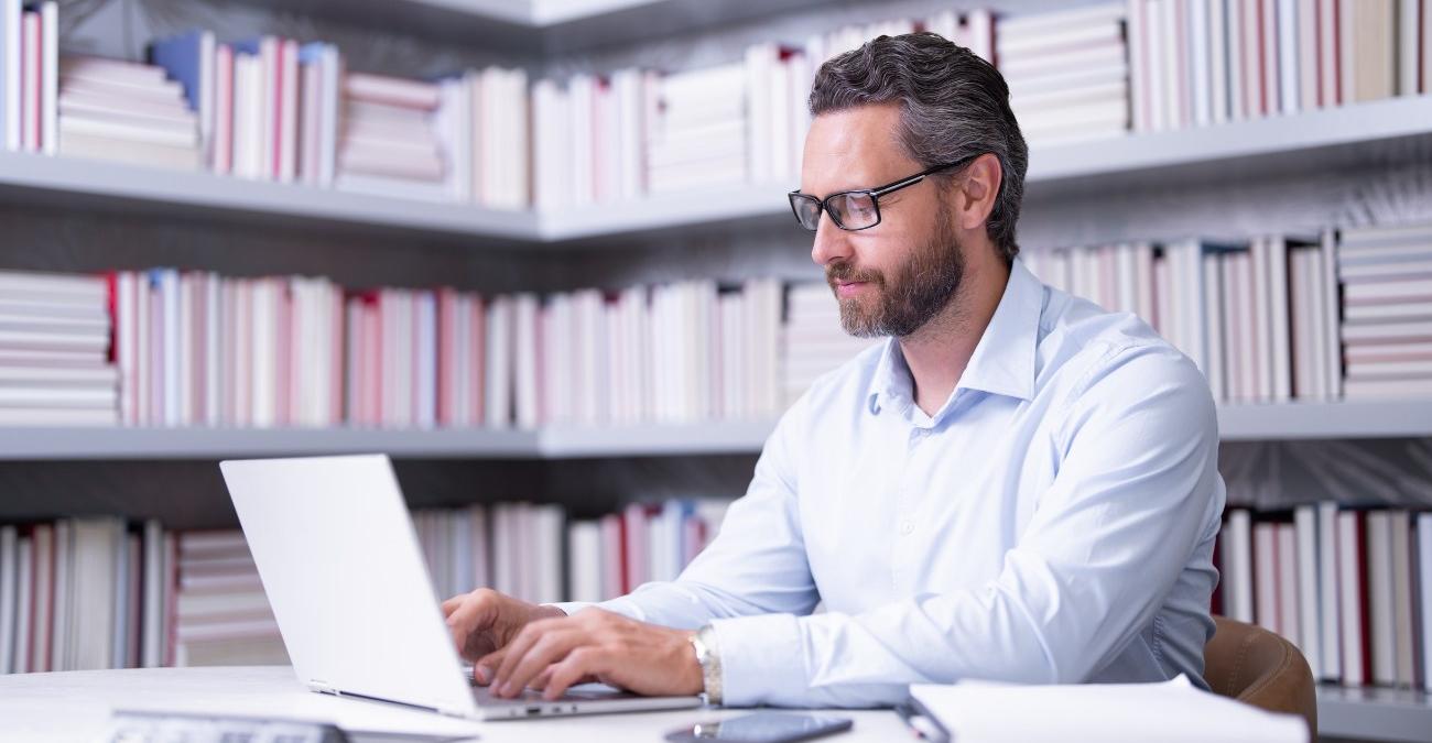 Ein selbstbewusster Geschäftsmann sitz mit seinem Notebook in einem modernen Büro mit Bücherregalen im Hintergrund – Sinnbild für Entscheider, die aus vielen Backup-Lösungen die für ihr Unternehmen passende auswählen.