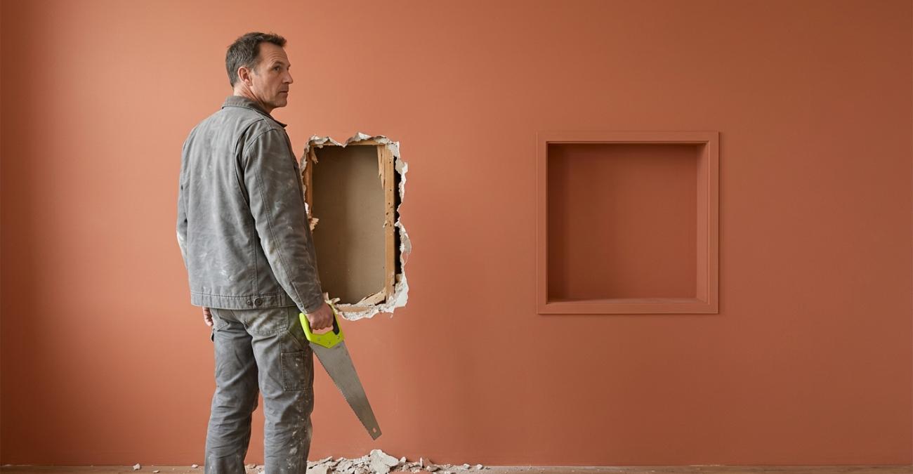 Ein Mann in staubiger Arbeitskleidung steht mit einer Handsäge vor einer terrakottafarbenen Wand, in die er eine grobe Öffnung gebrochen hat, während daneben bereits eine sauber gerahmte Öffnung sitzt. Sinnbild für eine barrierefreie Website, die nachträglich aufgestemmt statt von Anfang an eingeplant wird.