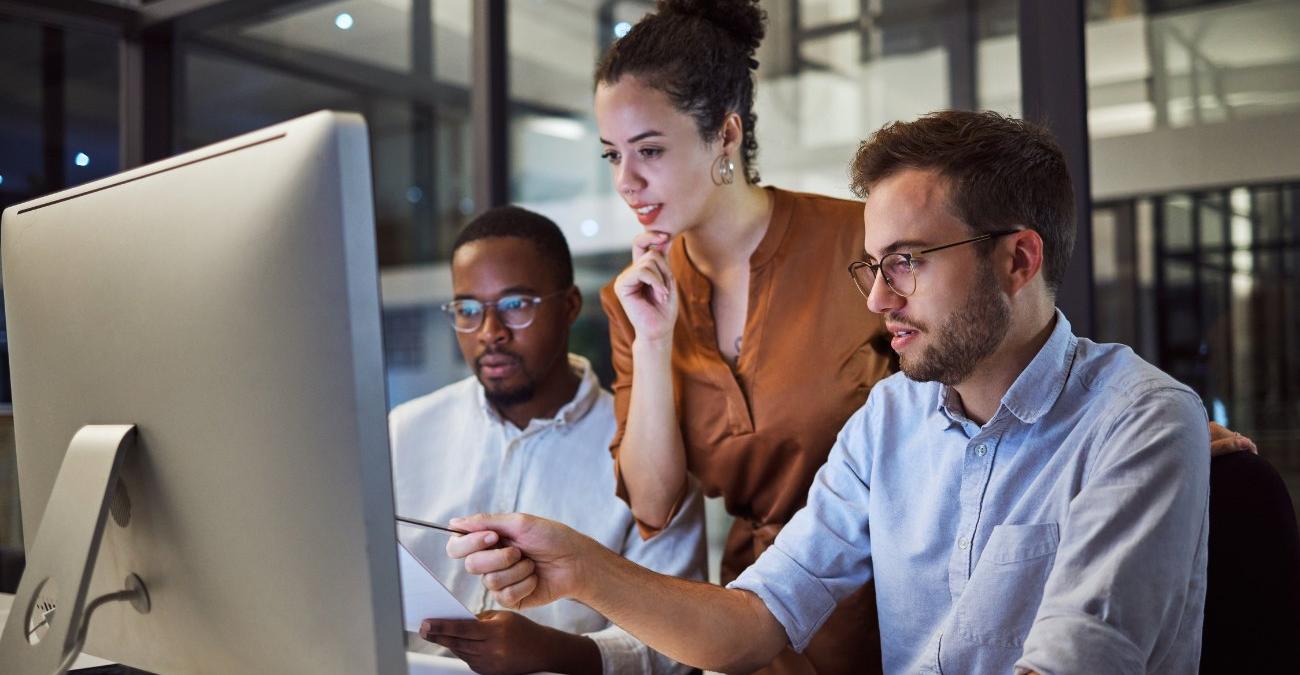 Ein helles Großraumbüro. Zwei Männer sitzen vor einem Rechner. Eine Frau steht zwischen ihnen. Sie ist die Chefin sieht sich fasziniert das barrierefreie Design ihrer Mitarbeiter aus der Grafikabteilung an.