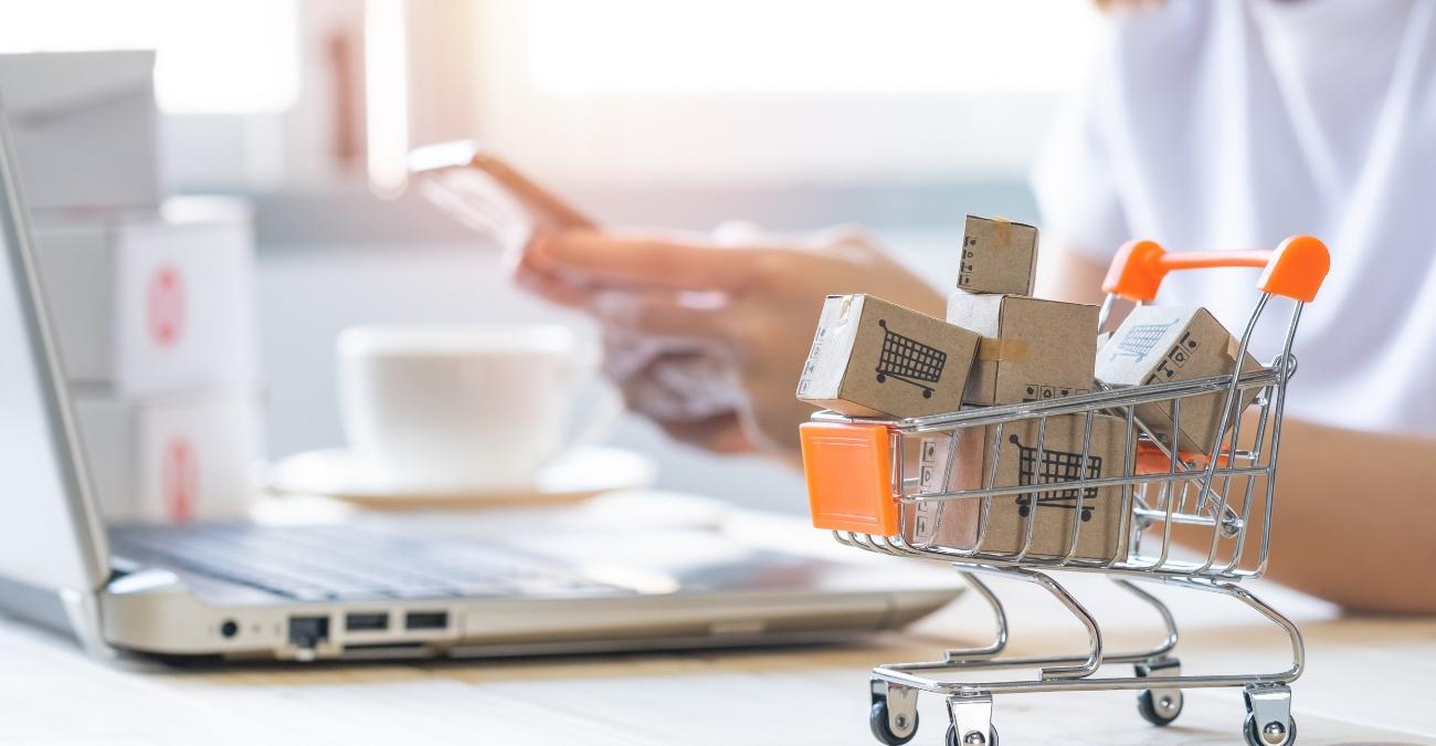 Eine Person sitzt am Laptop und hält ein Smartphone in der Hand, davor steht ein kleiner Einkaufswagen voller Paketkartons. Symbolbild für Barrierefreiheit im E-Commerce.