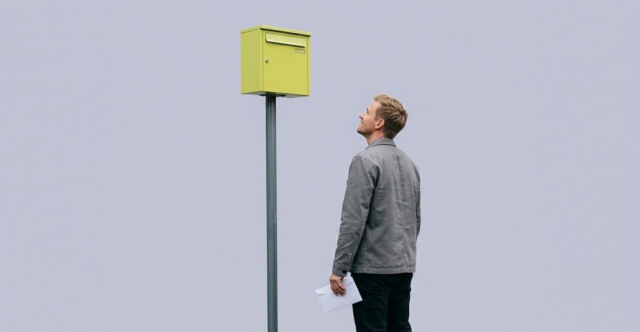 Ein Mann mit einem Briefumschlag in der Hand blickt zu einem gelbgrünen Briefkasten hinauf, der unerreichbar hoch an einem Pfahl sitzt. Sinnbild für Barrierefreiheit im Vergabeverfahren, wenn ein Verfahren formal offensteht und praktisch verschlossen bleibt.
