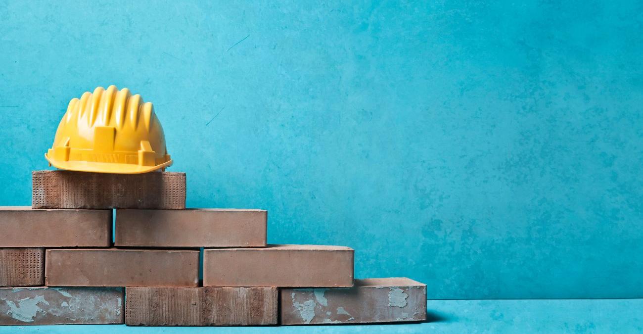 Blauer Hintergund. Im Vordergrund liegt ein Stapel Mauersteine, darauf ein gelber Bauerbeiterhelm, symbolisch für BSI-Grundschutz-Bausteine.
