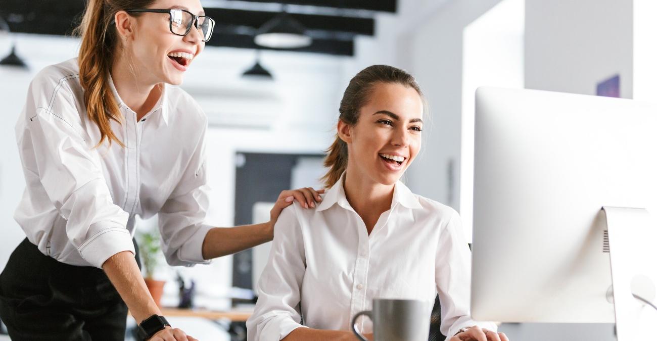 Zwei gut gelaunte Kolleginnen arbeiten im Büro gemeinsam am Computer und stimmen Maßnahmen für das Business Continuity Management ab.