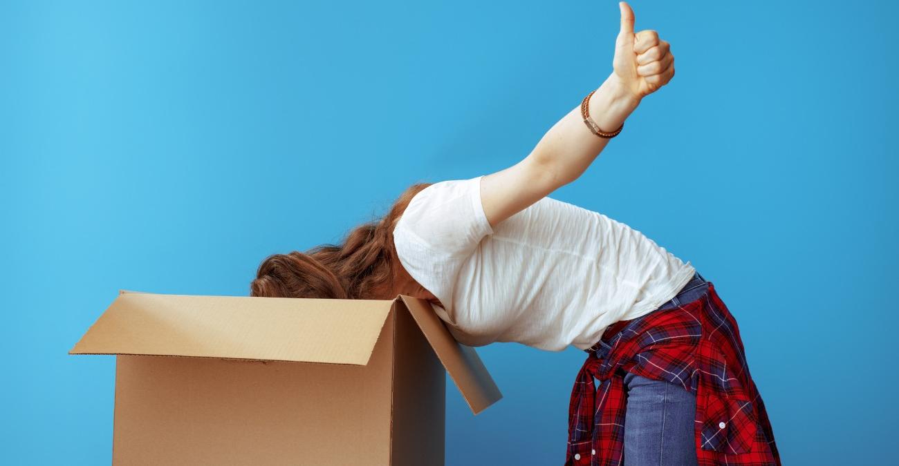 Einfarbiger, blauer Hintergrund. Eine junge Frau in legerem Outfit steckt ihren Kopf in einen großen Karton. Sie macht mit einer Hand eine Geste: Daumen hoch, symbolisch für eine gelungenes Datenmigration-Management.