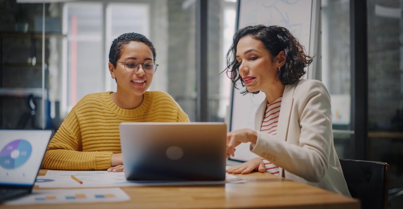 Die Ecke eine großen Büros. Die Chefin und ihre Assistentin sitzen am Tisch vor einem Notebook. Sie kümmern sich um die digitale Verwaltung.