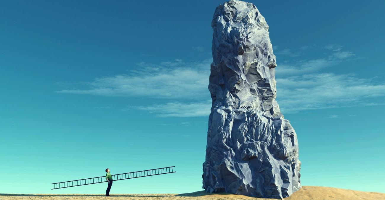 Ein großer Felsen vor blauem Himmel mitten in einer Wüstenlandschaft. Daneben steht - ganz klein - ein Mann mit einer langen Leiter. Das Bild versinnbildlicht, weleche einer Herausvorderung Patch Management für Konzerne sein kann.