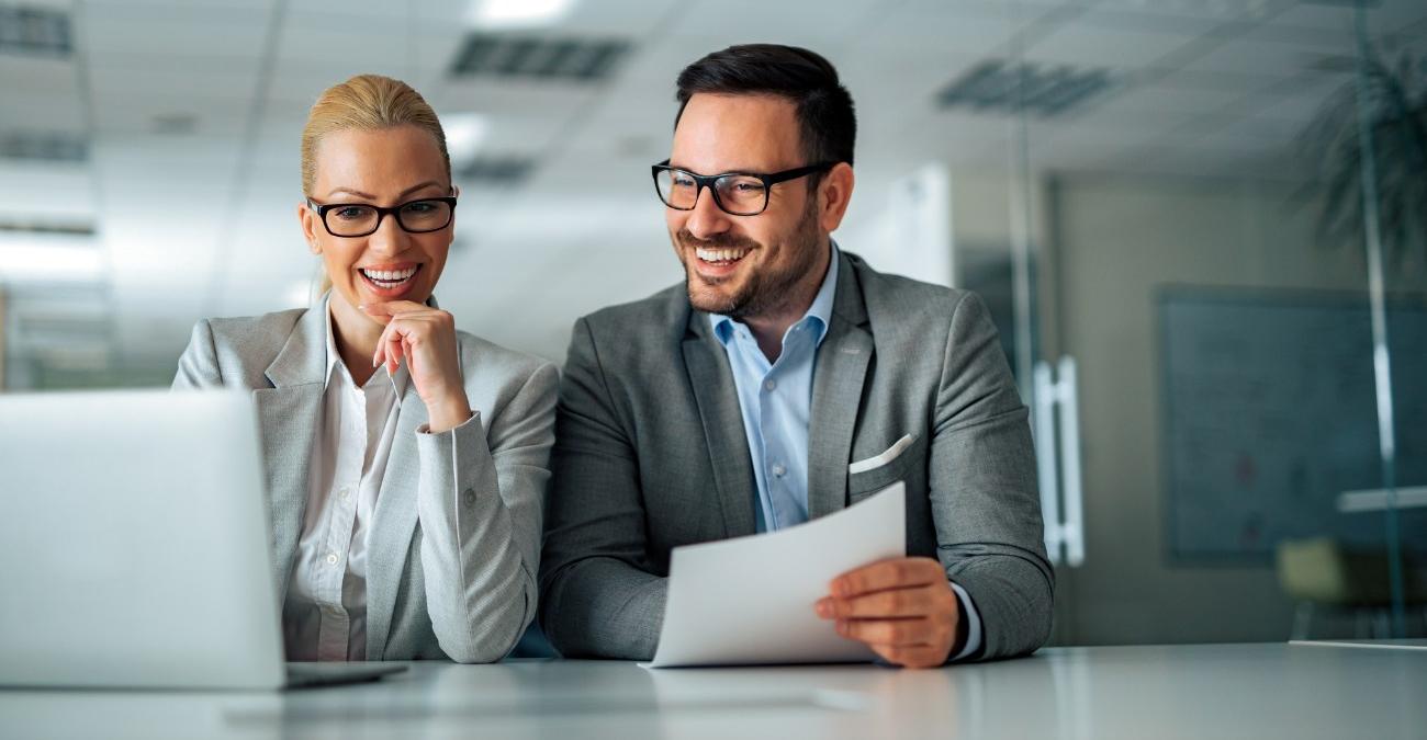 Eine Business-Frau und ein Geschäftsmann sitzen gut gelaunt in einem hellen Office. Sie schauen sich auf dem Notebook Daten zum ERP Customizing an.