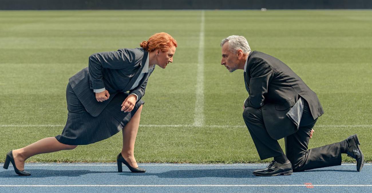 Eine Frau und ein Mann mittleren Alters im Business Outfit knien sich in einem leeren Sportstadion auf der Lauffläche in Startposition gegenüber, symbolisch für den Kampf zwischen ETL und ELT.