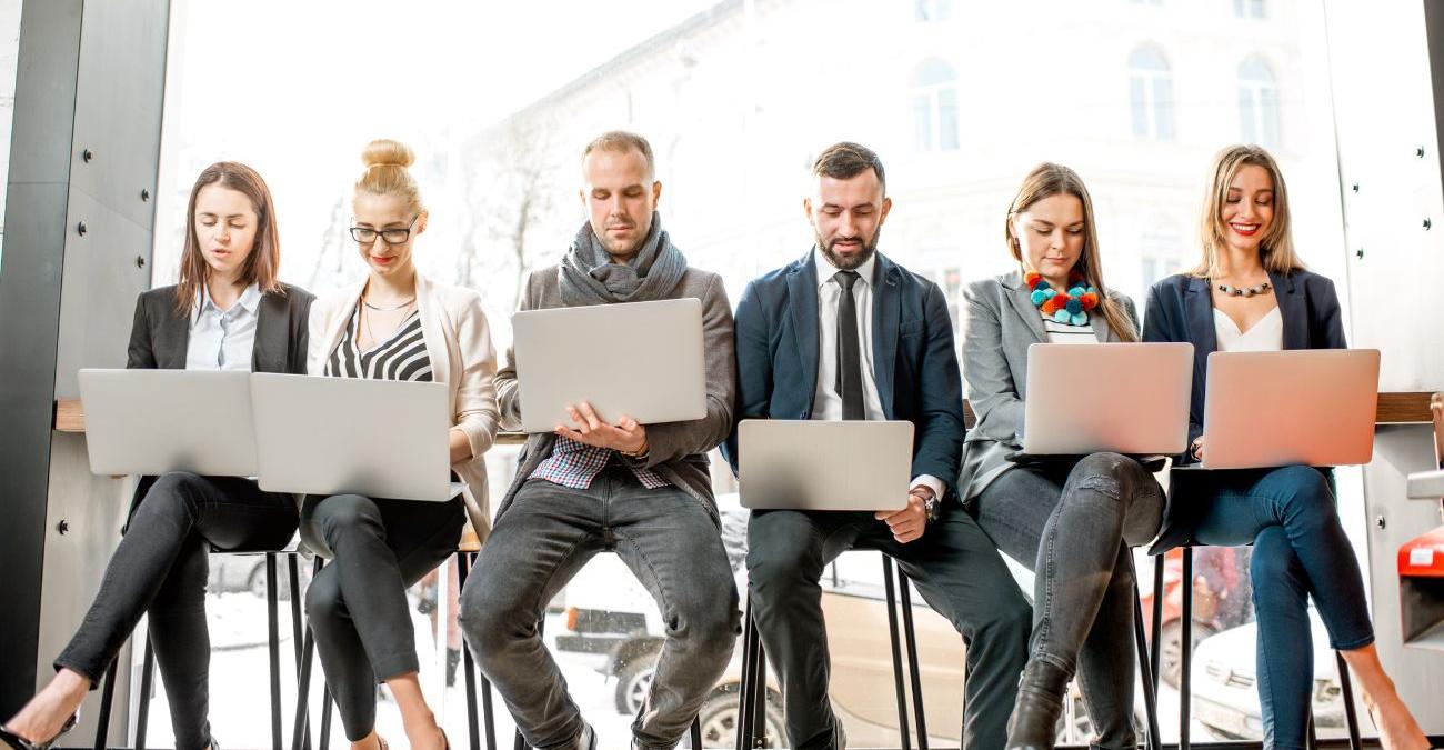Ein Wartezimmer. Mehrere Personen sitzen nebeneinander auf Barhockern. Alle arbeiten an ihren Notebooks. Sie profitieren von der Hochverfügbarkeit der IT-Infrastruktur.