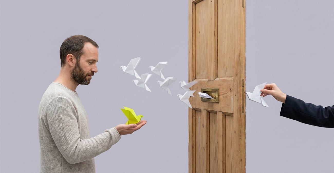 Ein Mann hält einen gelbgrünen Papiervogel in der offenen Hand, während weitere weiße Papiervögel aus dem Briefschlitz einer frei stehenden Holztür zu ihm fliegen und eine Hand auf der anderen Seite den nächsten hineinschiebt. Sinnbild für die Verbindung über die Zonengrenze nach IEC 62443-4-1, über die Daten nur in eine Richtung laufen.