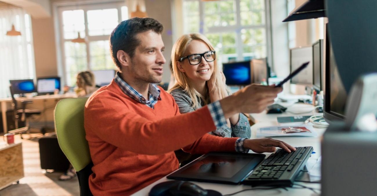 Das helle Office einer Softwareagentur. Auf einem halbrunden Tisch stehen diverse Bildschirme. Am vordersten Bildschirm sitzen eine junge Programmiererin und ihr Kollege. Sie sehen sich das neueste Produkt an, dass sie mit individueller App-Entwicklung erstellt haben.