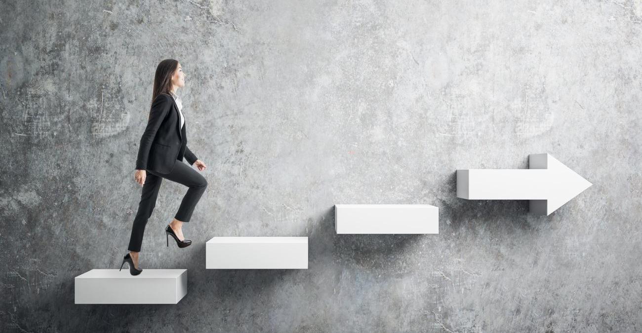 Eine graue, strukturierte Wand im Hintegrund. Davor weiße Stufen aus Beton, die aus der Wand zu ragen scheinen. Eine junge Frau im Business-Dress steigt diese Stufen hinauf. Sie folgt der Informationssicherheitsleitlinie.