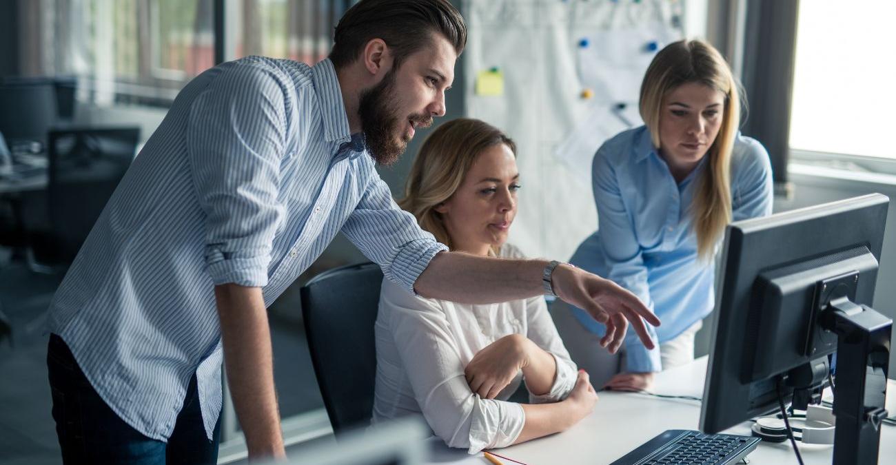 Drei Mitarbeitende eines Kleinunternehmens diskutieren vor dem Bildschirm eines Arbeitsplatzes über die bevorstehende ISO-22301-Zertifizierung ihres Business Continuity Management Systems.