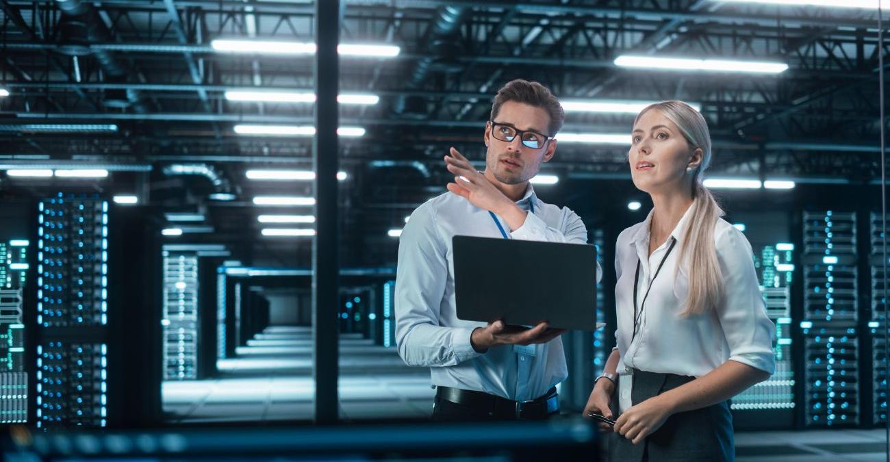 Ein Serverraum. Ein IT Mitarbeter steht neben einer Kollegin mit einem Notebook und erklärt den Unterschied zwischen ISO 27001 und IT-Sicherheit.