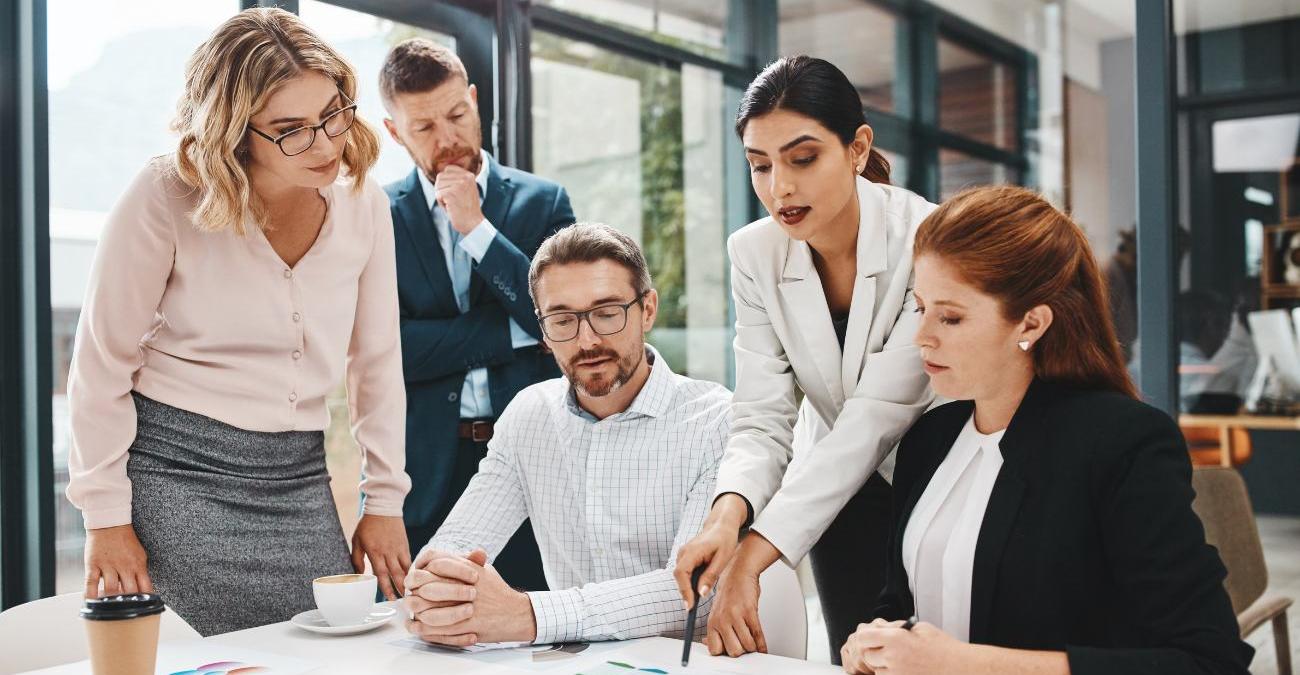 Mitarbeitende stehen im Büro um einen Tisch herum. Sie prüfen Dokumente zur Zertifizierung nach ISO-27001.