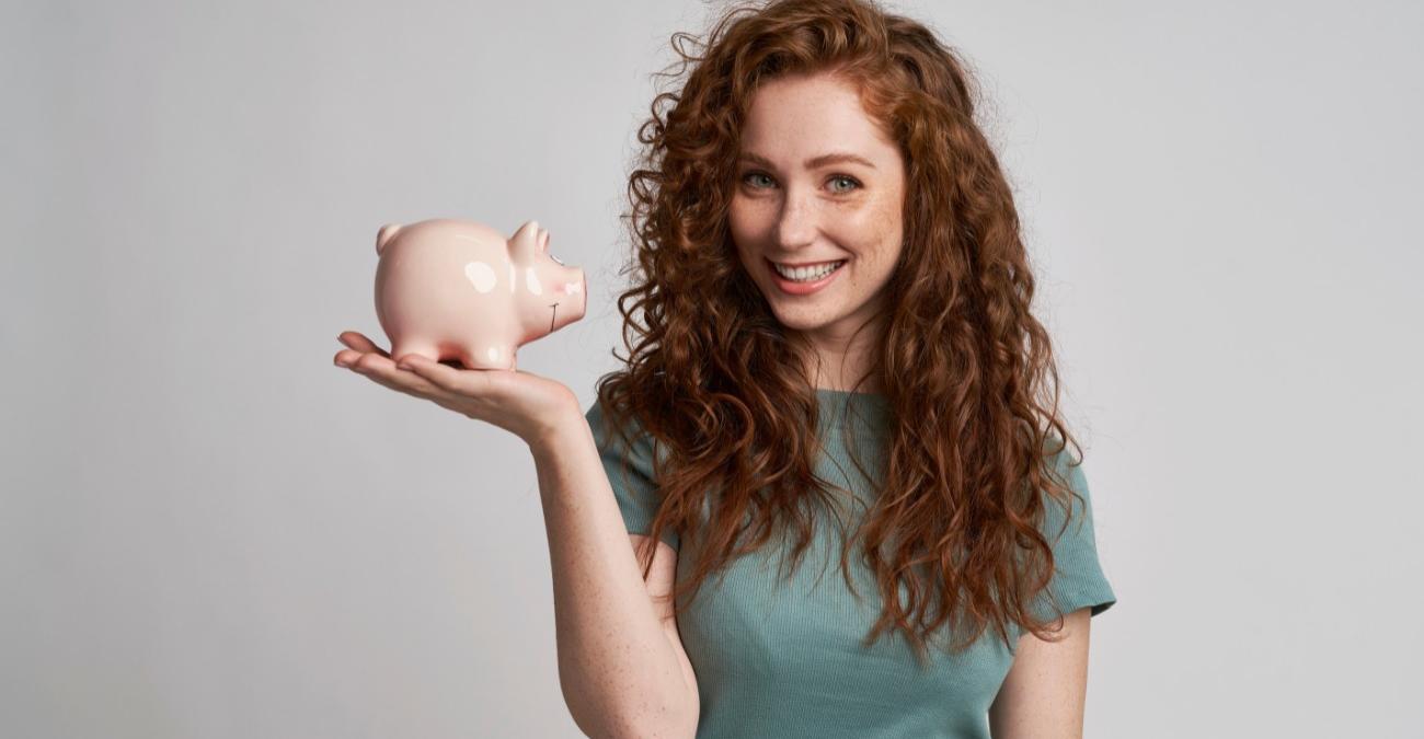 Neutraler, heller Hintergrund. Davor eine junge Frau mit grünem Shirt und wallenden roten Locken. Sie hält ein Sparschwein in der Hand und lächelt in die Kamera, weil sie bei ihrem IT-Dienstleister Kosten spart