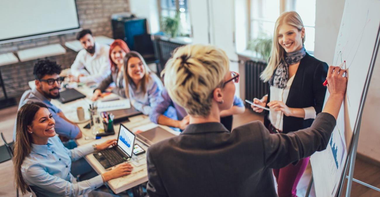 Ein langer Tisch in einem hellen Konferenzraum mit Backsteinwänden. Eine Frau steht vor einem Flipchart, um sie herum sitzen ihre Kollegen und Kolleginnen. Gut gelaunt besprechen sie die Maßnahmen zum IT-Grundschutz in der öffentlichen Verwaltung.