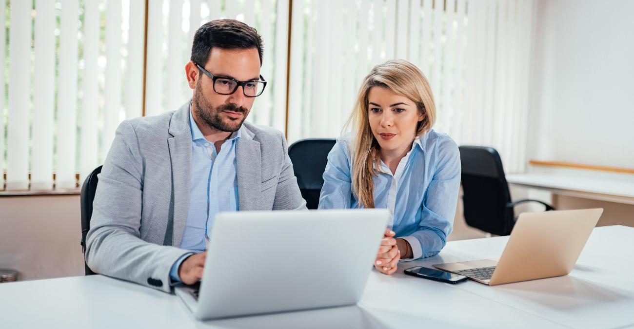 Ein helles Büro mit weißen Lamellen vor dem Fenster. Am großen Schreibtisch im Vordergrund sitzt die Abteilungsleiterin. Sie blickt auf den Notebook-Bildschirm des externen IT-Dienstleisters, der für sie eine IT-Konsolidierung planen soll.