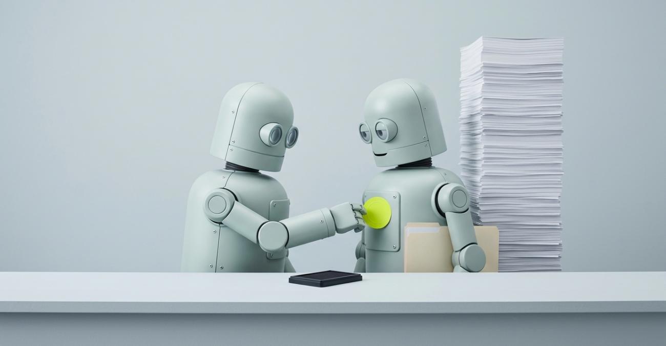 Zwei kleine graue Roboter stehen an einem Amtstresen, der linke drückt dem rechten einen gelbgrünen Aufkleber auf die Brustplatte, während hinter ihnen ein mannshoher Papierstapel aufragt. Ein Sinnbild für die KI-Beschaffung, bei der ein Amt durch den eigenen Namen zum Anbieter wird und damit den vollen Pflichtenkatalog übernimmt.
