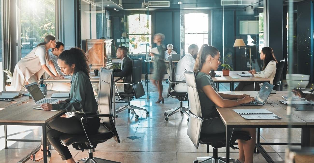 Ein Team aus Geschäftsmann, Geschäftsfrau und Mitarbeitenden arbeitet konzentriert in einem belebten Büro. Sie planen am Laptop und mit Unterlagen ihre Strategie zur KI-Compliance im Unternehmen.