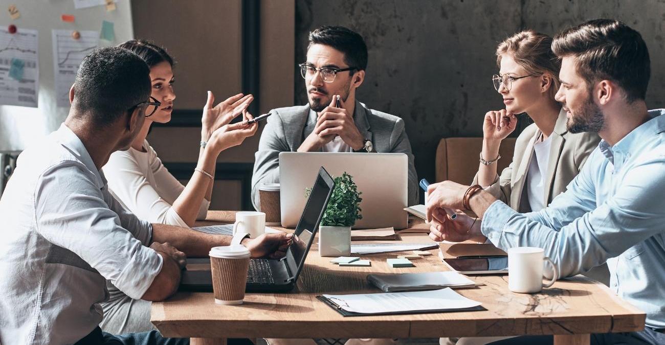 Eine Gruppe junger Menschen bespricht geschäftliche Themen in einem Meeting im Büro. Ein Symbolbild für IT-Compliance, bei der Teams gemeinsam regulatorische Vorgaben, Richtlinien und Sicherheitsanforderungen abstimmen.