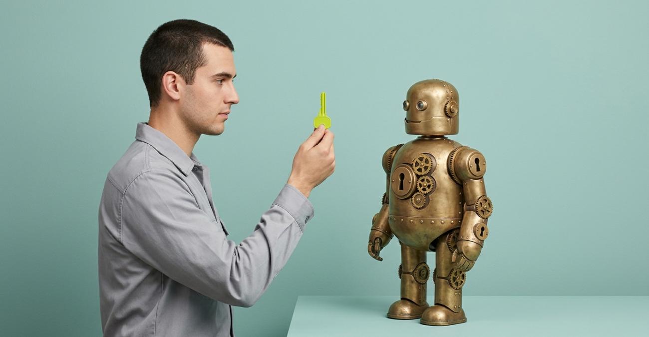 Ein junger Mann hält einem kleinen Messingroboter mit vier Schlüssellöchern einen gelbgrünen Schlüssel hin. Sinnbild für die KI-Readiness, die an vier Voraussetzungen zugleich hängt.