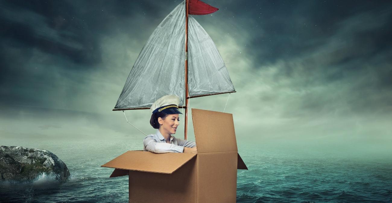 Eine rauhe See, wolkenbedeckter Himmel. Eine Frau mit Kapitänsmütze shippert wohlgelaunt in einem großen Karton mit weißem Segel durch die Fluten. Ein Symbolbild für das Ablösen der Legacy-Infrastruktur