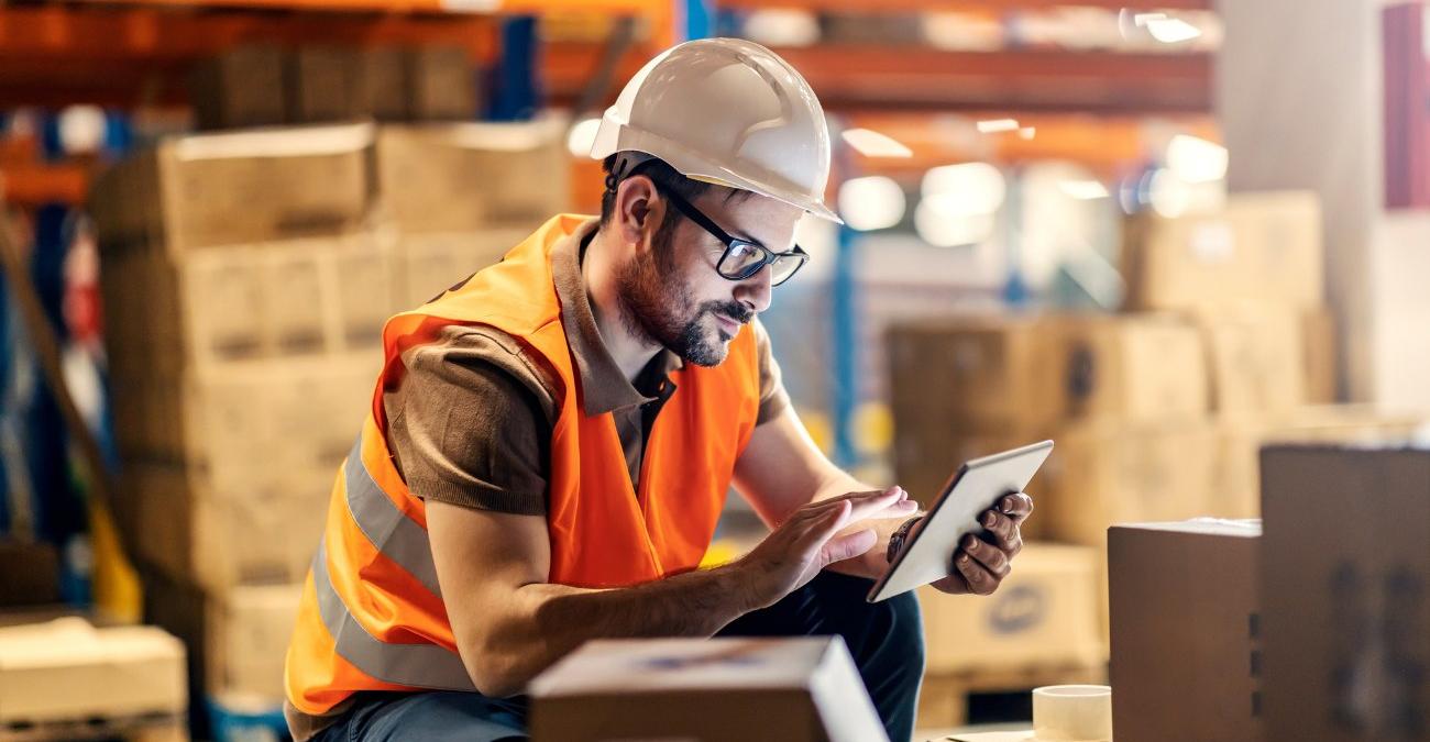 Ein Lagerarbeiter mit Warnweste und Helm prüft Bestände auf einem Tablet zwischen Kartons im Lager, ein Symbolbild für den praktischen Einsatz von Logistiksoftware.