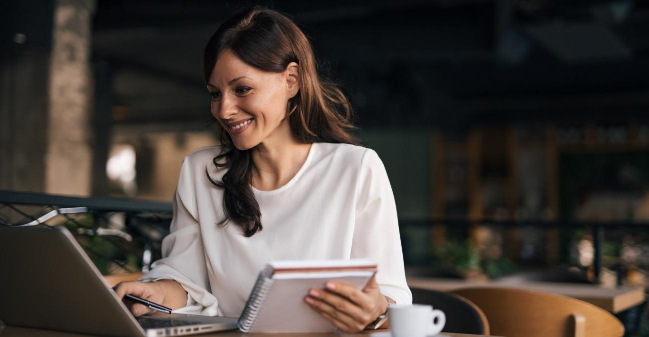 Eine gut gelaunte Frau mit Notizblock am Laptop in einem Restaurant, die dank reibungsloser Datenflüsse über eine Middleware ortsunabhängig auf alle Unternehmenssysteme zugreift.