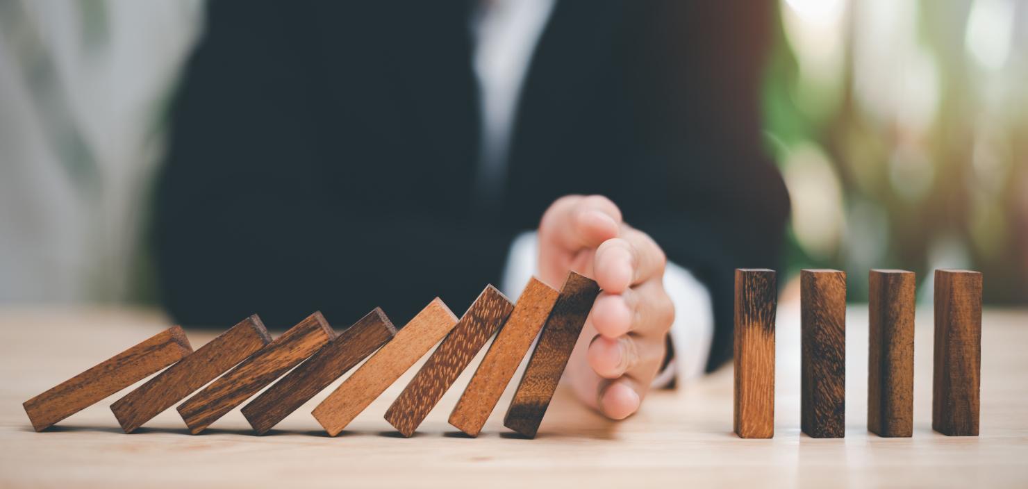 Dominosteine aus Holz. Sie fallen. Ein Mann stoppt sie mit der Hand. Ein Symbolbild für NIS2-Lieferkettensicherheit.