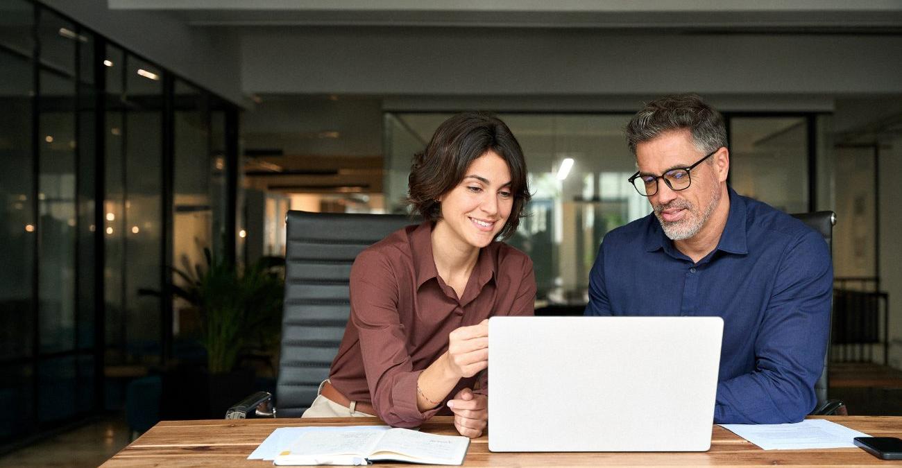 Ein großes Office. Ein IT-Support-Mitarbeiter sitzt am Notebook neben der Chefin. Er berät sie zum Patch Management.