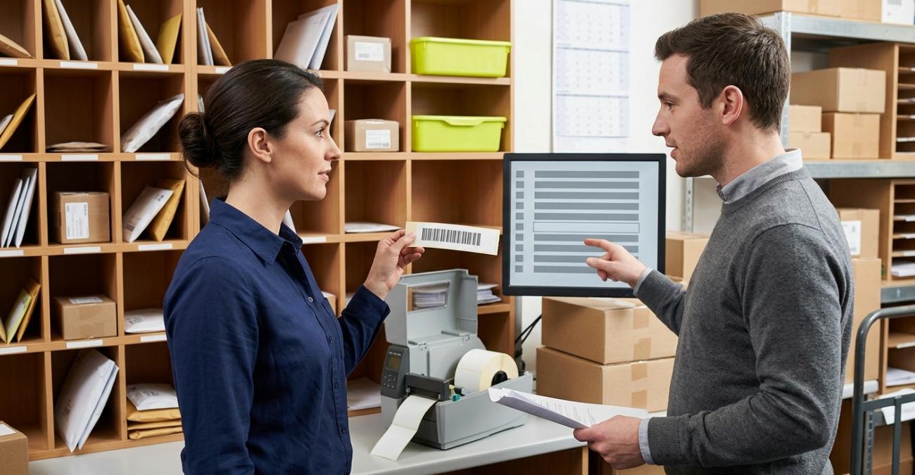 Zwei Personen in einer Poststelle mit Fächerregal, Kartons und Etikettendrucker: links hält eine Frau ein frisch gedrucktes Etikett hoch, rechts zeigt ein Mann auf einen Monitor, auf dem eine Liste gleichmäßiger Balkenzeilen eine leere Zeile enthält. Sinnbild dafür, dass eine Peppol-ID erst durch den Eintrag im Verzeichnis erreichbar wird.