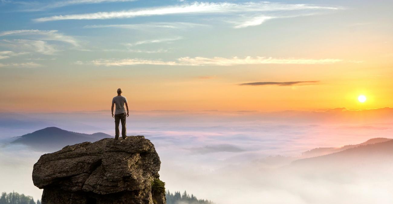 Ein Mann im Freizeit-Look steht auf einem großen Felsen und blickt über die Wolken in den Sonnenaufgang. Ein Symbolbild für PHP-Legacy-Modernisierung.