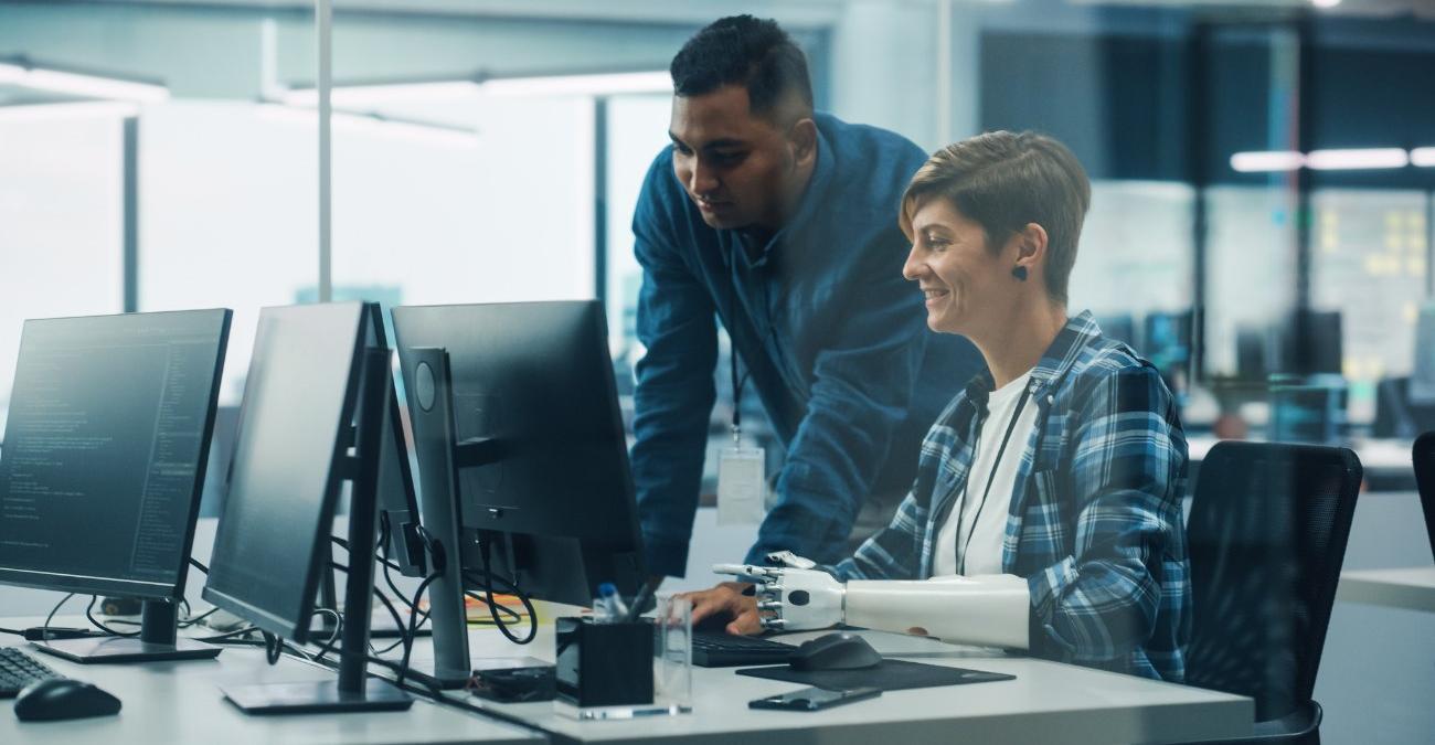 Teamwork in einem inklusiven Büro: Ein Mitarbeiter steht am digitalen Arbeitsplatz einer Kollegin mit Armprotese. Ihre Software ist barrierefrei.