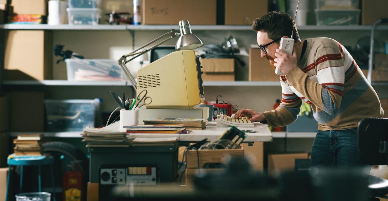 Ein altes Lager. Ein Mann mit einem uralten Funktelefon steht vor einem alten Bildschrim. Hier sind Hardware und Software end-of-life.