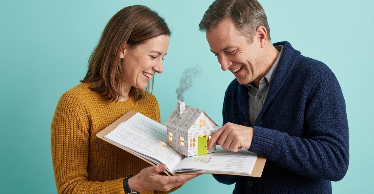 Eine Frau und ein Mann beugen sich lachend über eine aufgeschlagene Dokumentenmappe, aus deren Seiten sich ein kleines Haus mit beleuchteten Fenstern und rauchendem Schornstein aufgefaltet hat, der Mann öffnet mit dem Finger die gelbgrüne Haustür. Sinnbild für Software Escrow, bei dem aus hinterlegten Unterlagen im Ernstfall wieder eine laufende Anwendung wird.