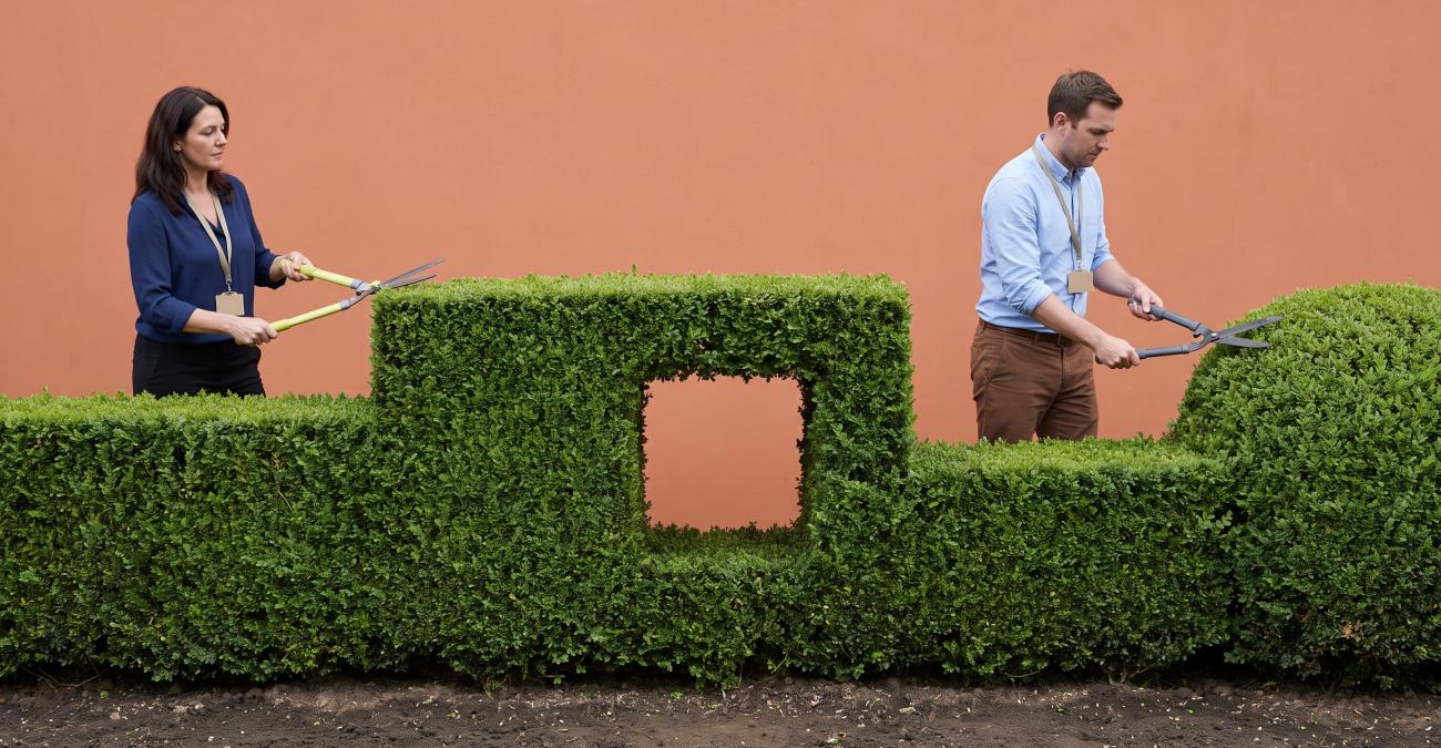 Zwei Personen in Bürokleidung mit Ausweisband schneiden jeweils ihre Hälfte einer durchgehenden Hecke, in deren Mitte ein exakt rechteckiges Loch offen bleibt, um das sich niemand kümmert. Sinnbild für Software in der öffentlichen Verwaltung, deren Lücken zwischen den Systemen liegen.