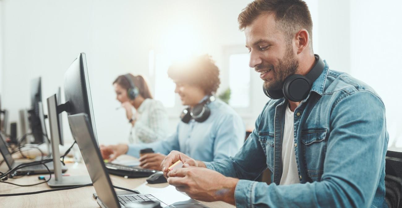 Mehrere IT-Servicemitarbeiter sitzen nebeneinander an Service-Desks. Sie sind gut gelaunt und kümmern sich um die Softwarewartung.