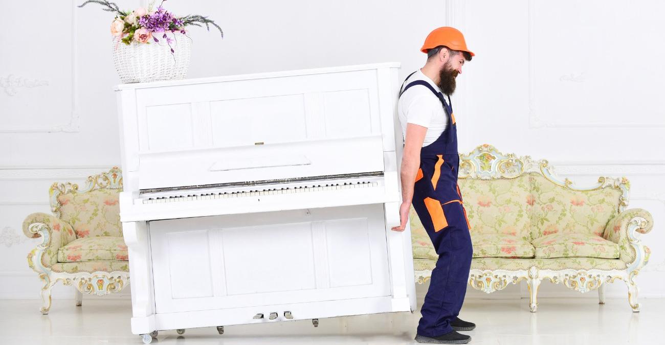 Ein weißer Raum mit einem Biedermeier-Sofa im Hintergrund. Davor wuchtet ein Bauarbeiter mit einem orangen Helm ein weißes Konzertpiano durch den Raum. Ein Symbolbild für Symfony-Migration.
