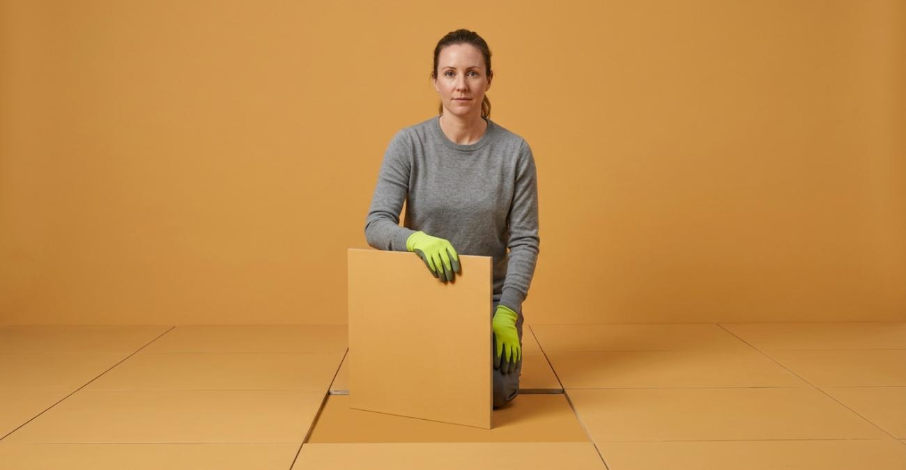 Eine Frau mit gelbgrünen Arbeitshandschuhen kniet auf einem ockerfarbenen Fliesenboden und hält eine herausgenommene Bodenplatte aufrecht vor sich. Sinnbild für den Third Level Support, der eine Störung bis unter die Oberfläche verfolgt.