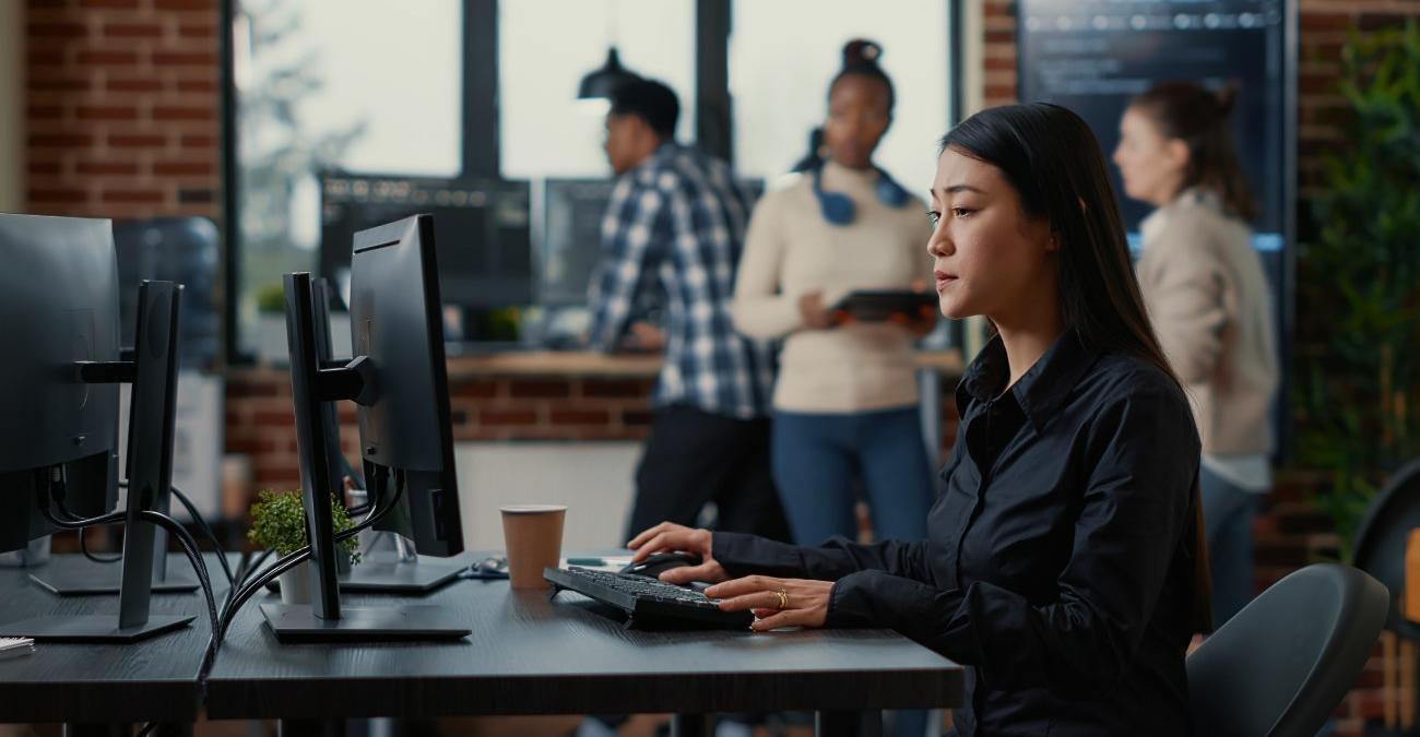 Eine junge Entwicklerin sitzt im großen, modernen Office der Typo3-Agentur an ihrem Rechner und arbeitet konzentriert. Im Hintergrund sind unscharf ihre Mitarbeiter zu sehen.