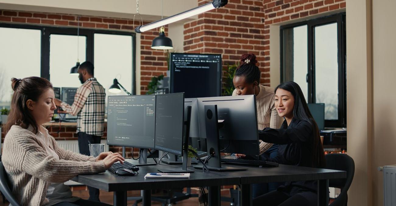 Eine moderne Webdesign-Agentur. Programmiererinnen sitzen an ihren PCs an einem großen Schreibtisch und kümmern sich um die Typo3-Entwicklung.