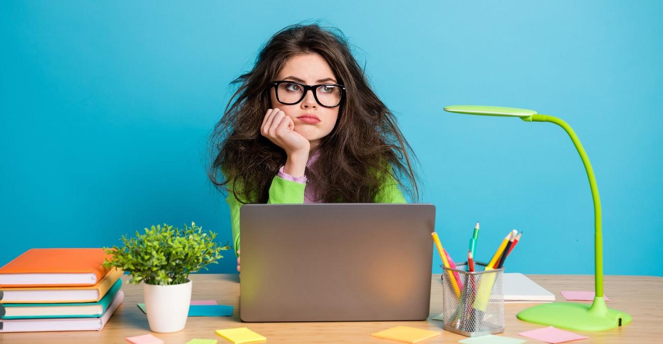 Hellblauer Hintergrund. Bunte Schreibutensilien auf dem Schreibtisch. Eine junge Frau mit zerzaustem Haar sitzt am Notebook. Sie blickt nachdenklich ins Weite und denkt über Typo3 Migration nach.