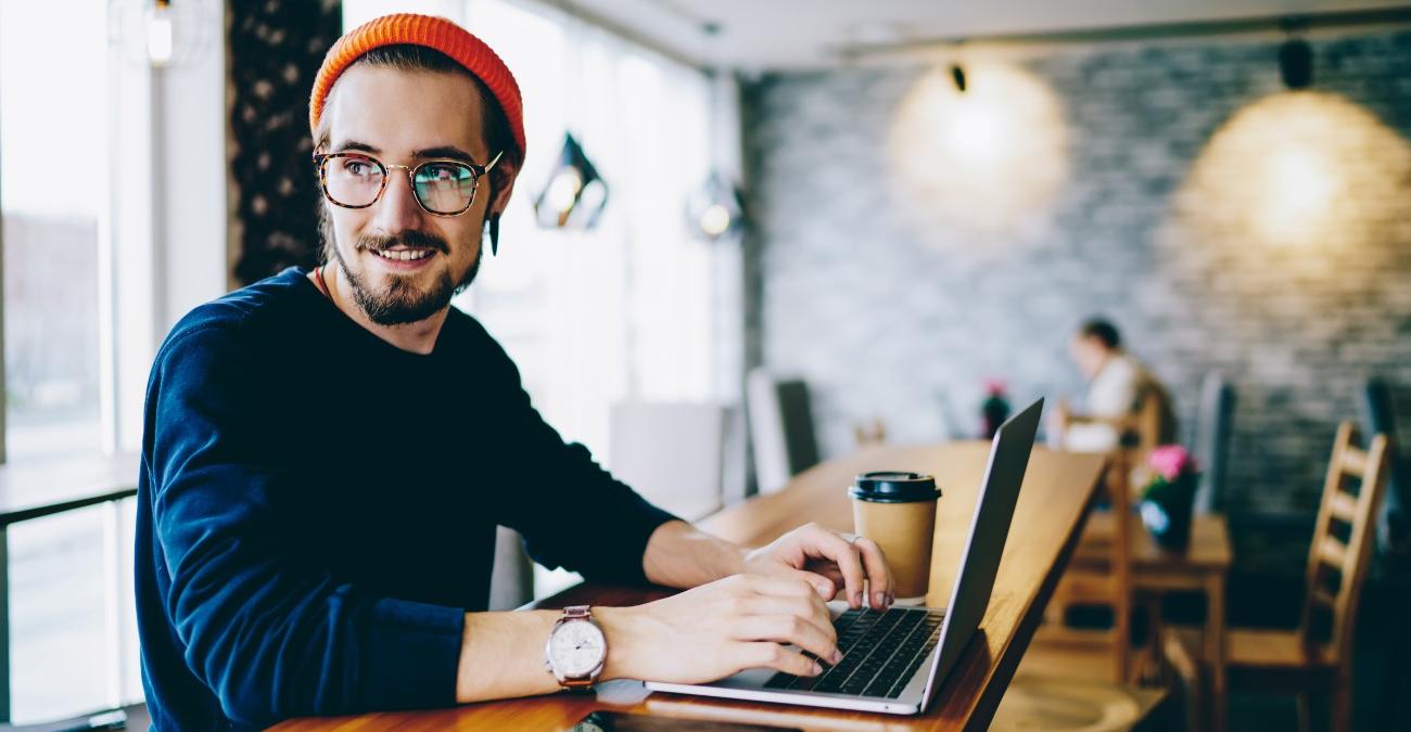 Ein junger, hipper Mann sitzt mit seinem Notebook im Café. Er blickt zur Seite und redet mit jemandem, während sein Typo3 Update läuft.