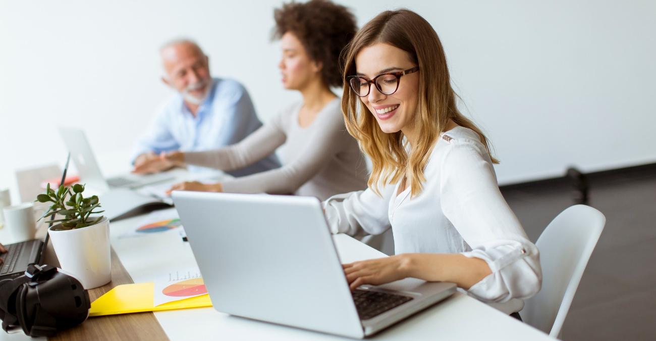 Eine junge Web-Entwicklerin macht gut gelaunt ihre Type3-Wartung am Notebook im Büro. Ihre Kollegen unterhalten sich im Hintergrund.