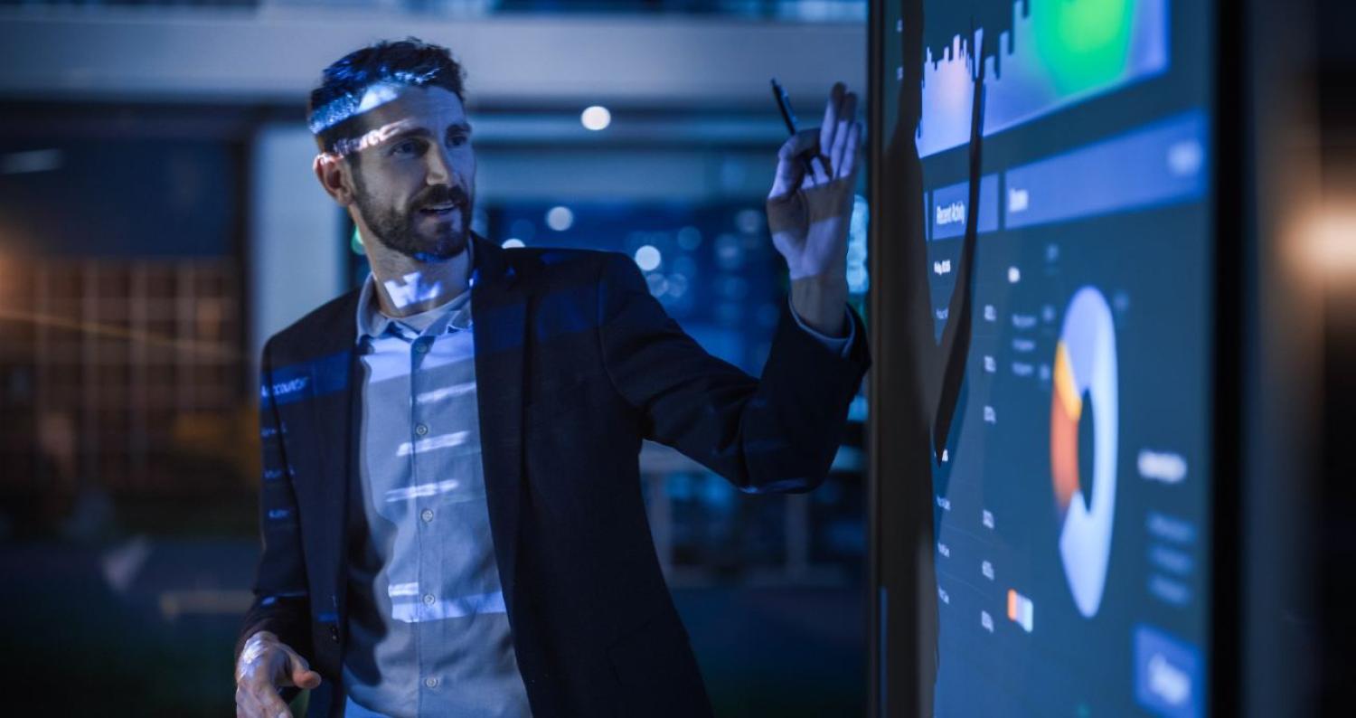 Ein Angestellter steht bei gedämpftem Licht vor einem Clipboard. Er analysiert die Auswertung der Verwaltungsprogramme seiner Organisation.